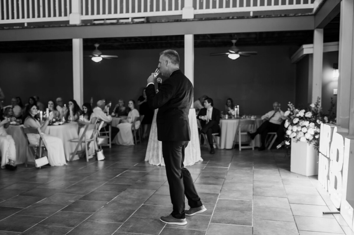 A man in a suit speaks into a microphone at a wedding reception, with guests seated at decorated tables in the background. The setting has tiled floors and a balcony above. The image is in black and white.