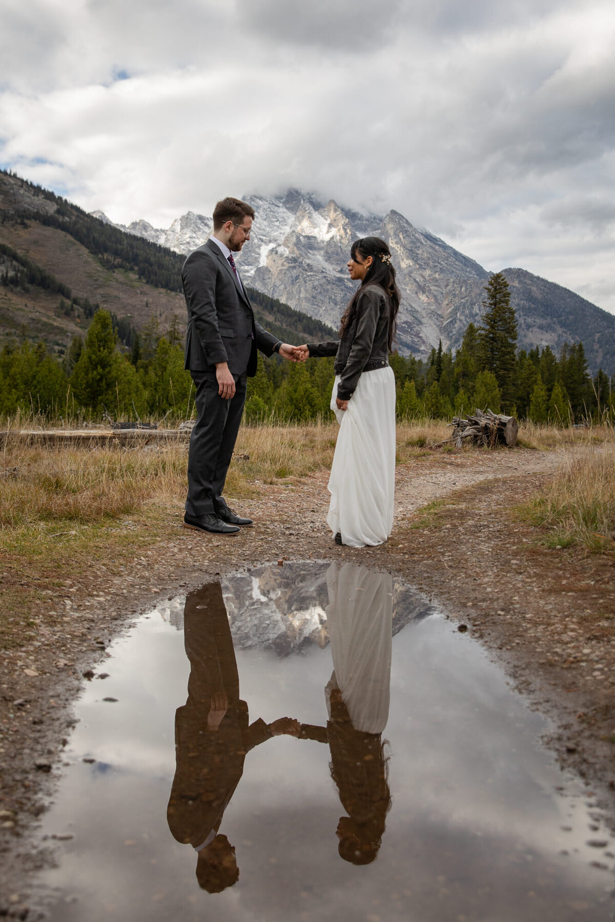 A bride and groom stand holding hands while their Wyoming elopement photographer uses a puddle to capture their reflection.