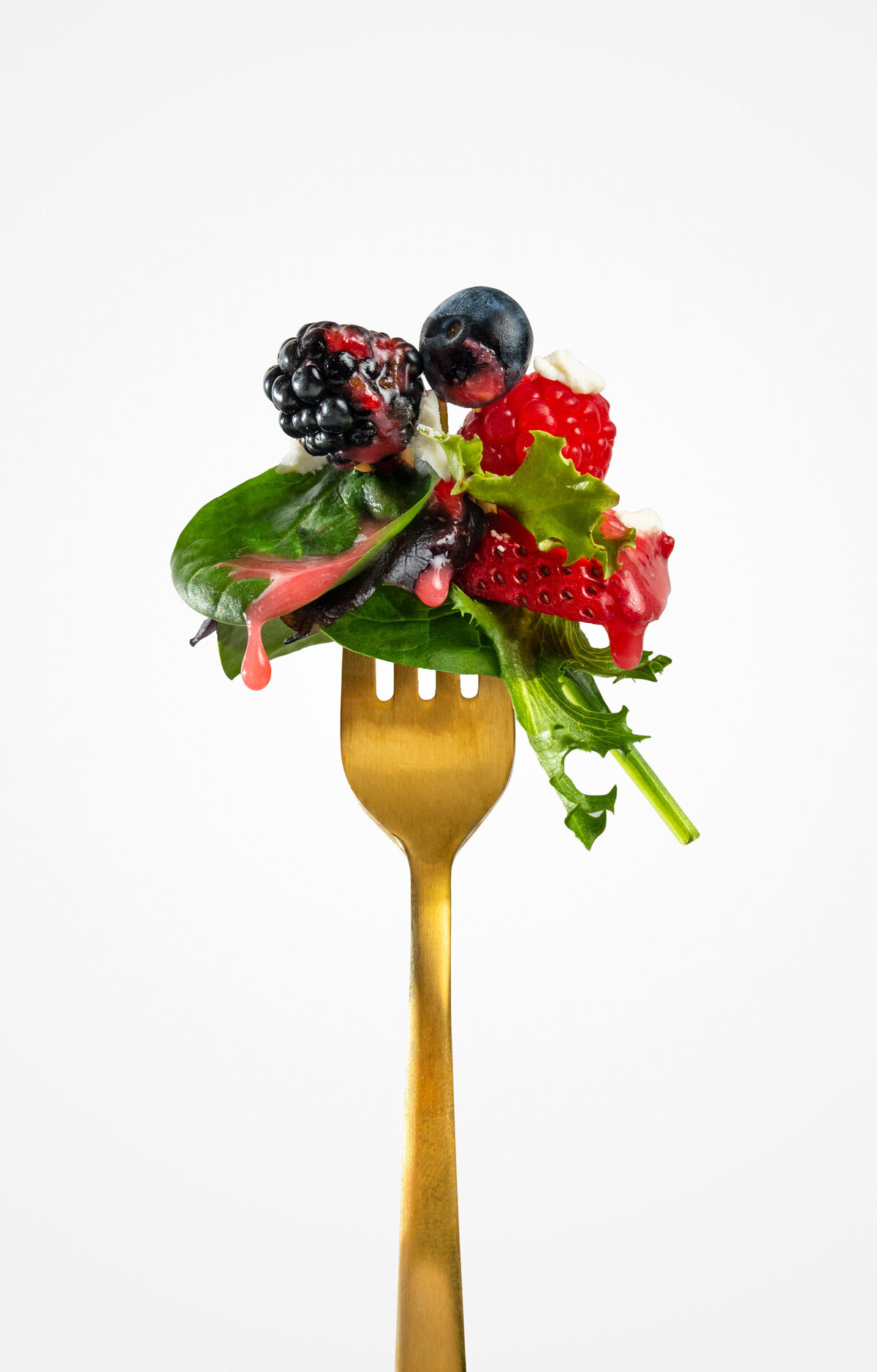 A spring berry salad with homemade raspberry vinaigrette dripping off of spinach leaf, on a golden fork.