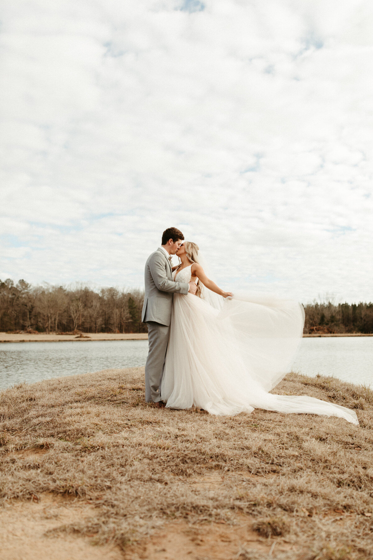 Bride waving tulle wedding dress as her groom is holding and kissing her with a lake behind them