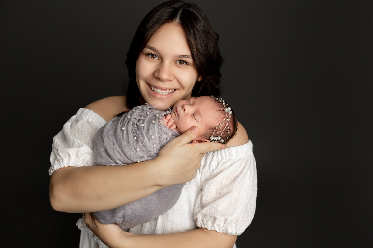 New mother lovingly cradling her peacefully sleeping newborn daughter wrapped in a pearl-adorned gray swaddle, with a matching pearl headband, against a dark studio backdrop. Captured by Inland Empire newborn photographer Kaitlyn Dawn Photography.