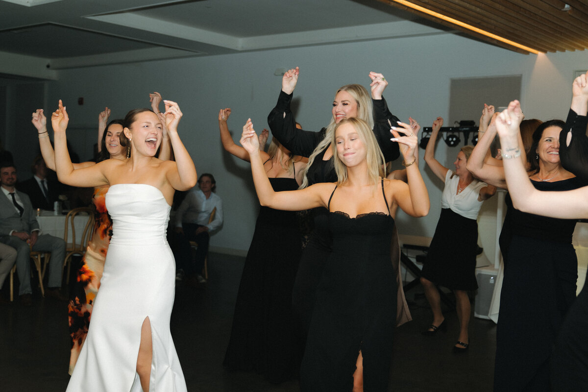 A bride in a white strapless, fitted wedding dress smiles as she snaps while glancing over her left shoulder at a women in a black bridesmaid dress also snapping her fingers and smiling with her eyes closed. Bridesmaids in formal, black, floor length dresses also raise their arms up and snap smiling at each other. 