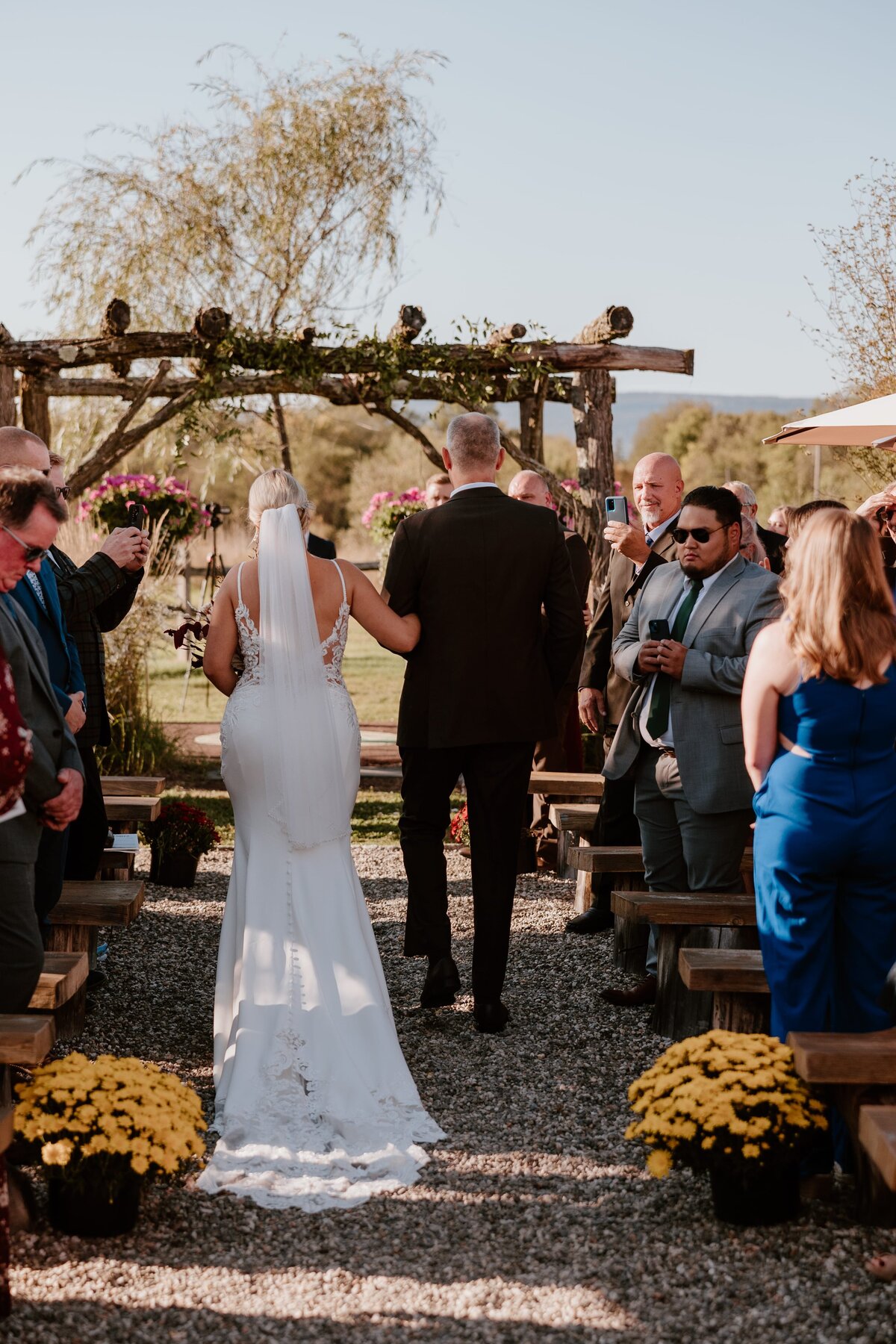 A bride walks down the aisle with her father at Lippincott Manor, toward a rustic wooden arch decorated with greenery and flowers. The bride wears a lace-back gown with a long veil, and guests stand and capture the moment on their phones. The outdoor setting, under a clear blue sky, provides a beautiful backdrop for this emotional moment. Yellow flowers and wooden benches line the aisle, adding to the warm and inviting atmosphere.