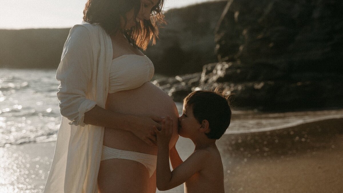 L'enfant embrasse le ventre rond de sa mère, en maillot, face à la mer. Lumière dorée. Séance bord de mer par Kamescop, photographe et vidéaste.