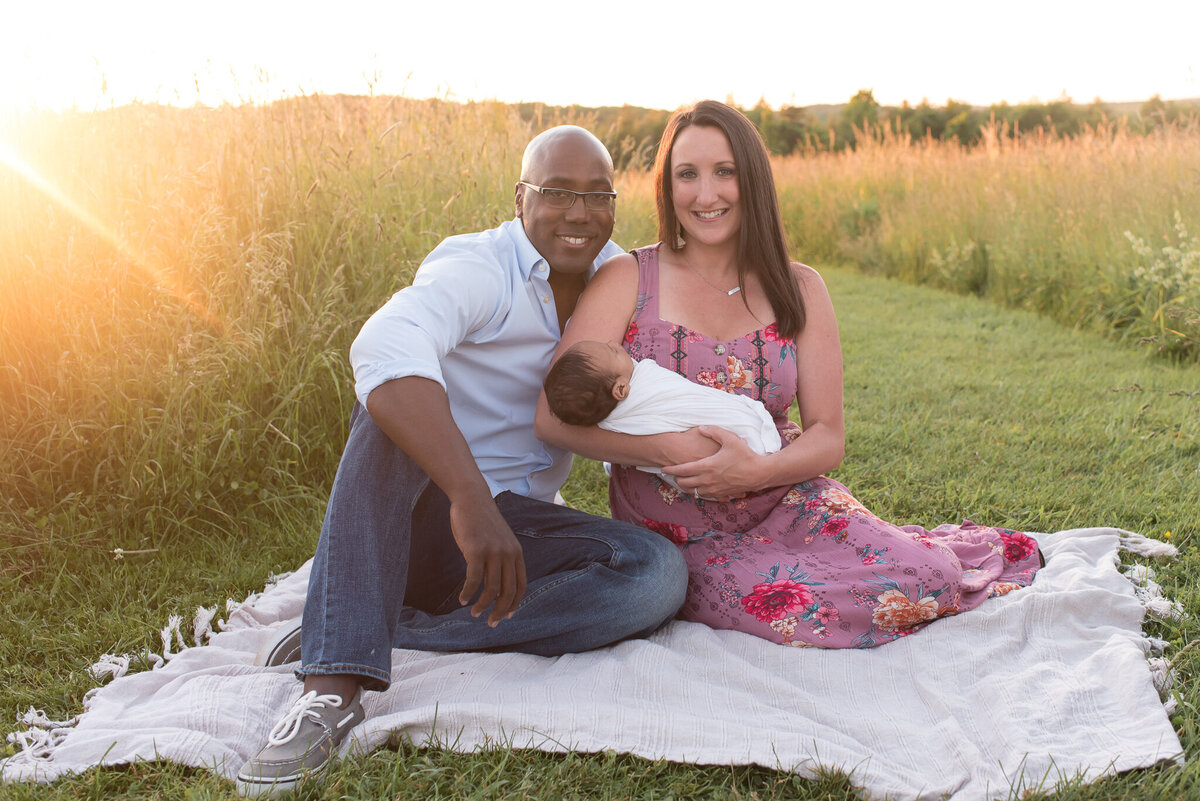 A man and a woman are sitting on a blanket, holding their infant and smiling at the camera with the sun setting in the field behind them