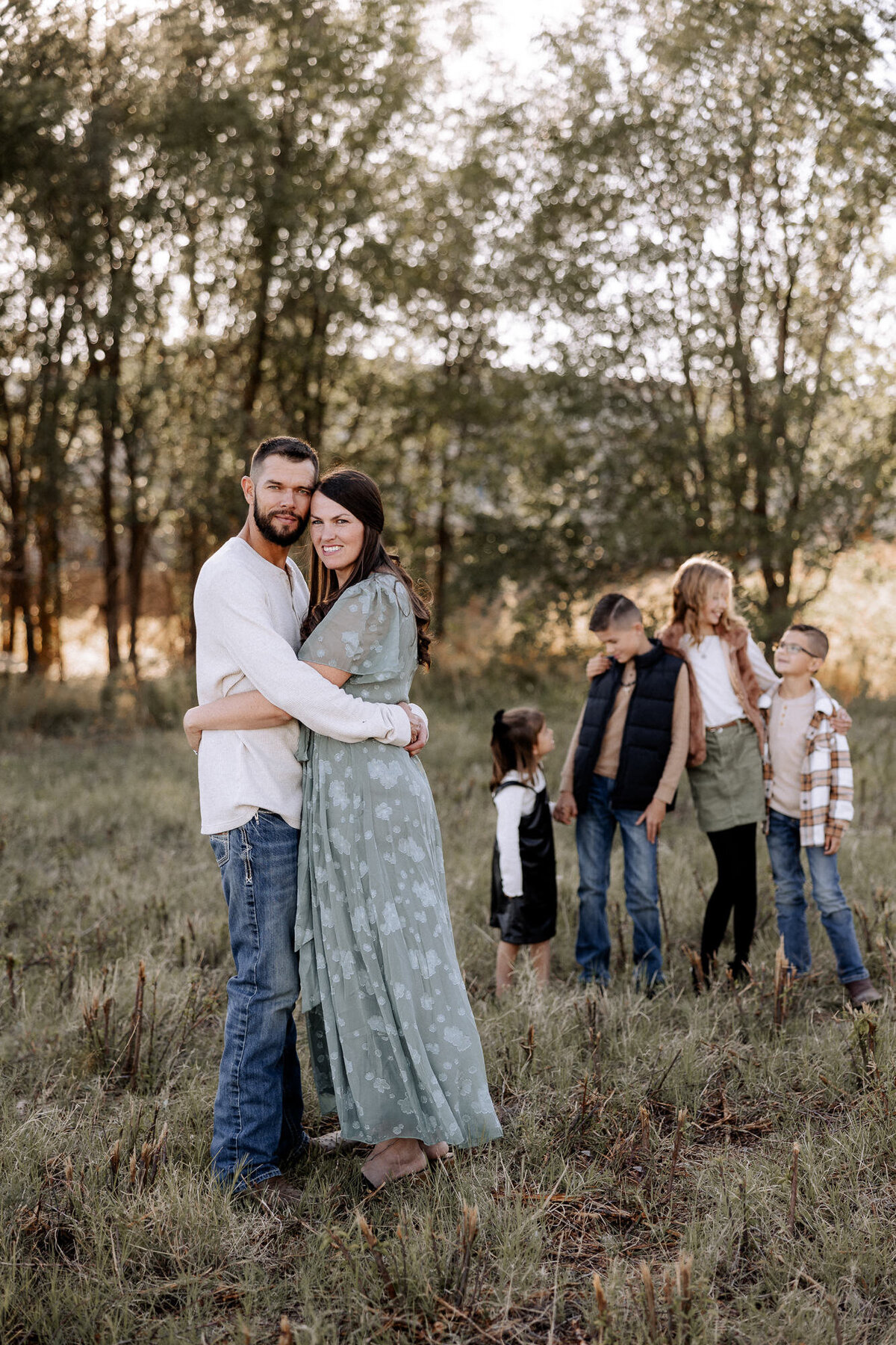 A heartfelt family portrait set in a sunlit field, featuring a loving couple embracing while their children share playful moments in the background. This warm and natural setting captures the beauty of family bonds and the joy of being together. Photographed by Mary Wiebe Photography in Lubbock, Texas.