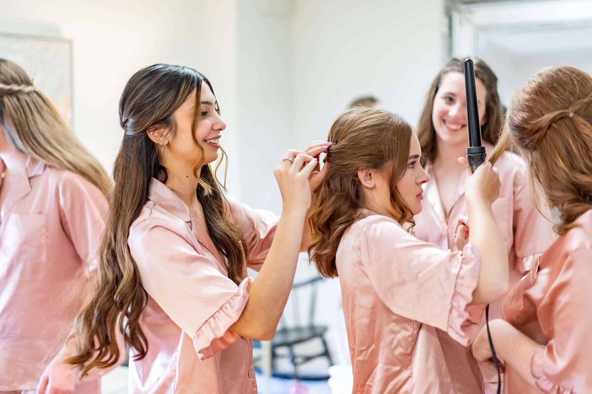 A group of young women in matching pink outfits are styling each other’s hair, smiling and preparing together in a bright bridal suite. One woman uses a curling iron while another helps pin a friends hair.