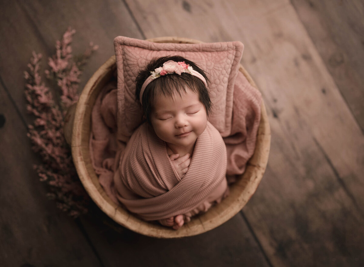 Aerial image of Menifee newborn baby photoshoot. Baby girl is wrapped in a dusty pink and placed in a bowl. Her toes and fingers are peeking out of the swaddle. She is sleeping peacefully. Captured by best Menifee newborn photographer Bonny Lynn Photography