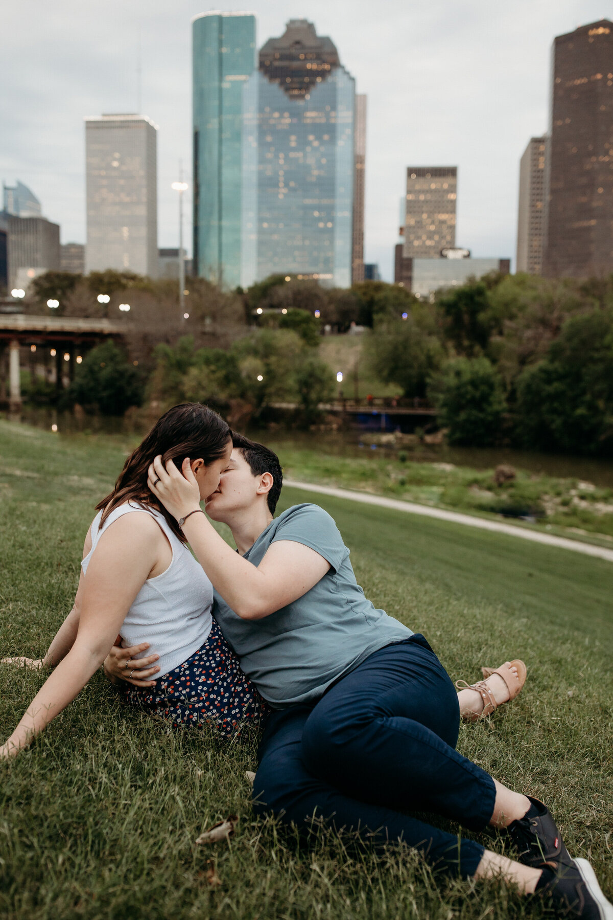 A couple sharing a kiss on a grassy field with a city skyline in the background during dusk