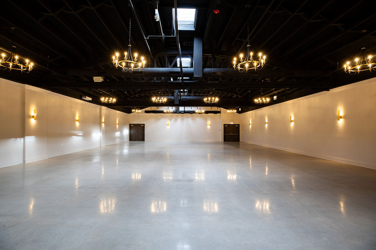 Spacious ceremony, reception or event space, with high industrial ceilings, polished floors, neutral walls and details, and ample natural light streaming through skylights and all around sconces and chandeliers, ready to be set up for an event