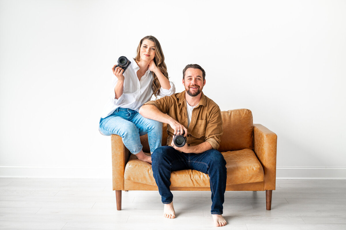 Reg and Kala holding their camera's sitting on a large caramel couch. Kala is sitting on the arm of the chair and Reg is sitting on the chair.