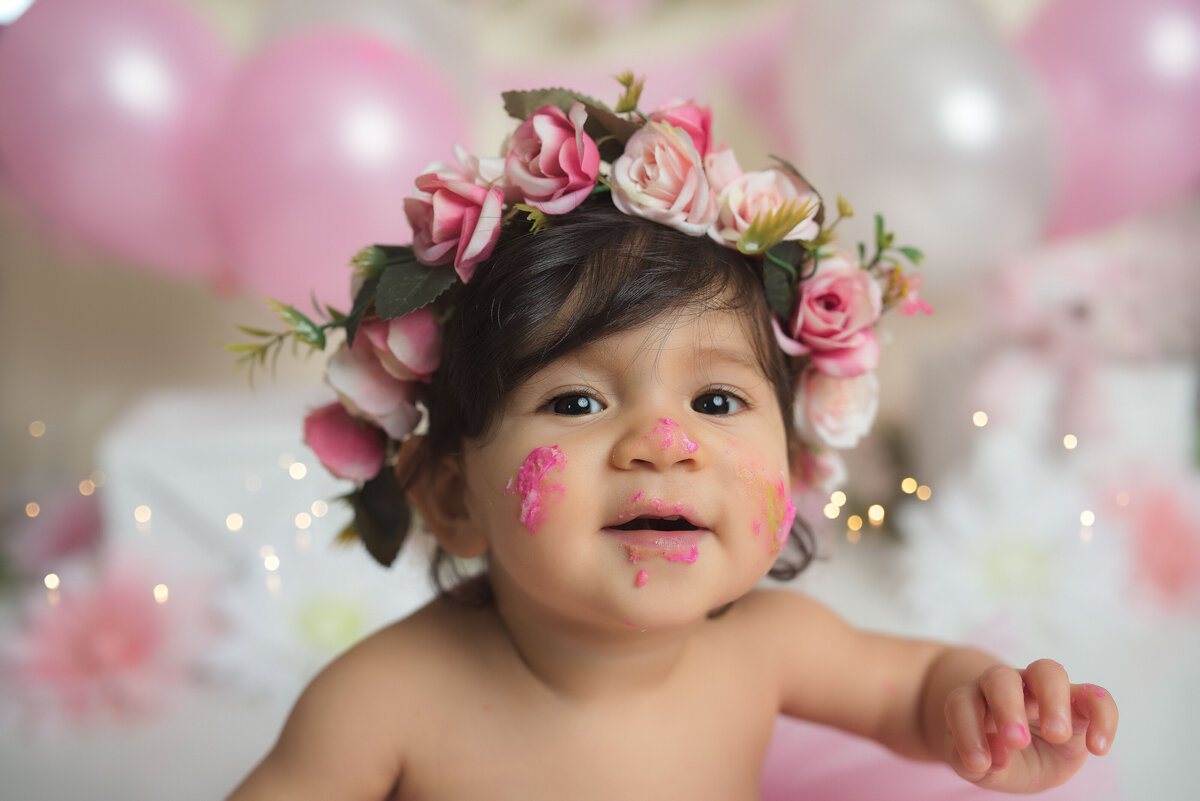 One year old baby girl with pink icing on her nose and cheeks smiling and wearing a pink floral crown with pink and white balloons in background and twinkle lights