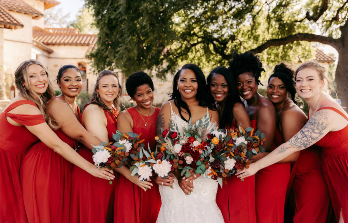 A stunning bride surrounded by her wedding party in vibrant red gowns, each with soft curls and glowing makeup. Perfect example of wedding party hair and makeup services available at our San Antonio salon, including expert bridal hairdressers and flawless airbrush makeup.