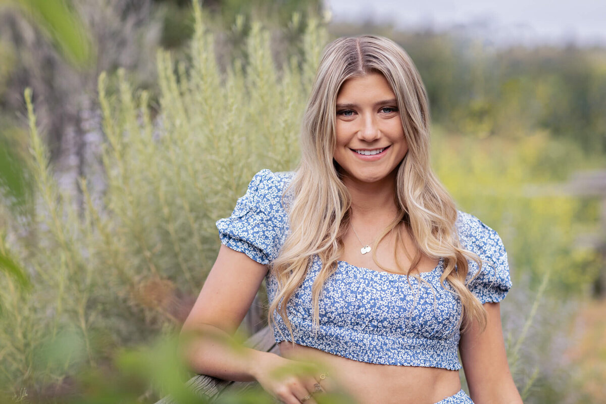 A young woman with long blonde hair smiles while sitting outdoors. She wears a blue floral crop top with puffed sleeves. The background features green foliage and a soft focus on distant trees.