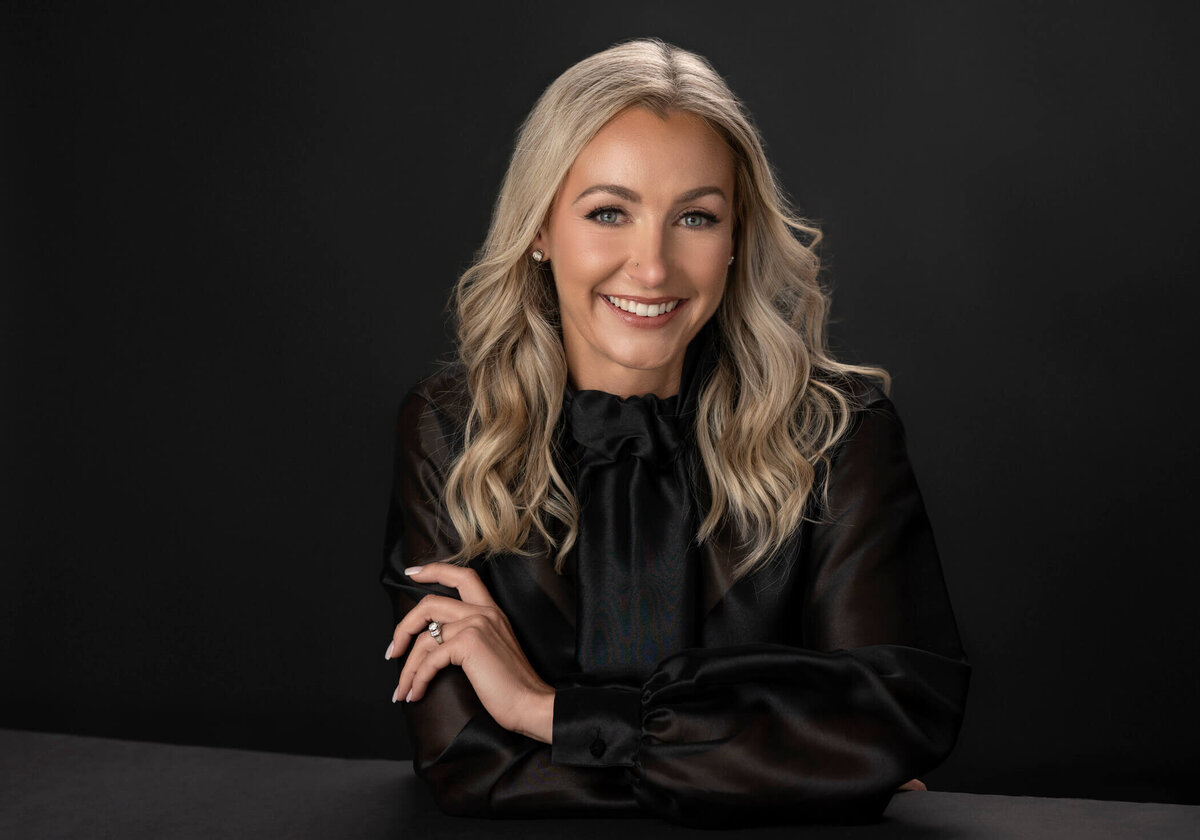 A blonde woman with wavy hair, wearing a black blouse with sheer sleeves, smiling in a dark, moody setting.