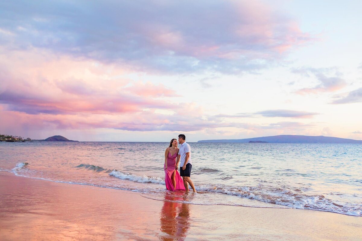 Happy couple laughs at sunset in Wailea while holding hands and walking on the beach
