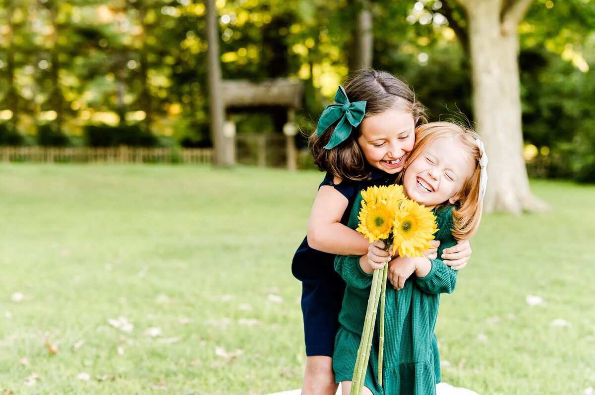 Girls laughing with sunflowers as they play during  family session in Geneva, IL.