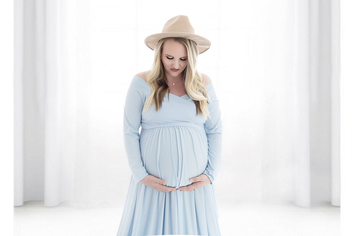 A mom to be in a blue maternity gown smiles down at her bump while standing in a studio window