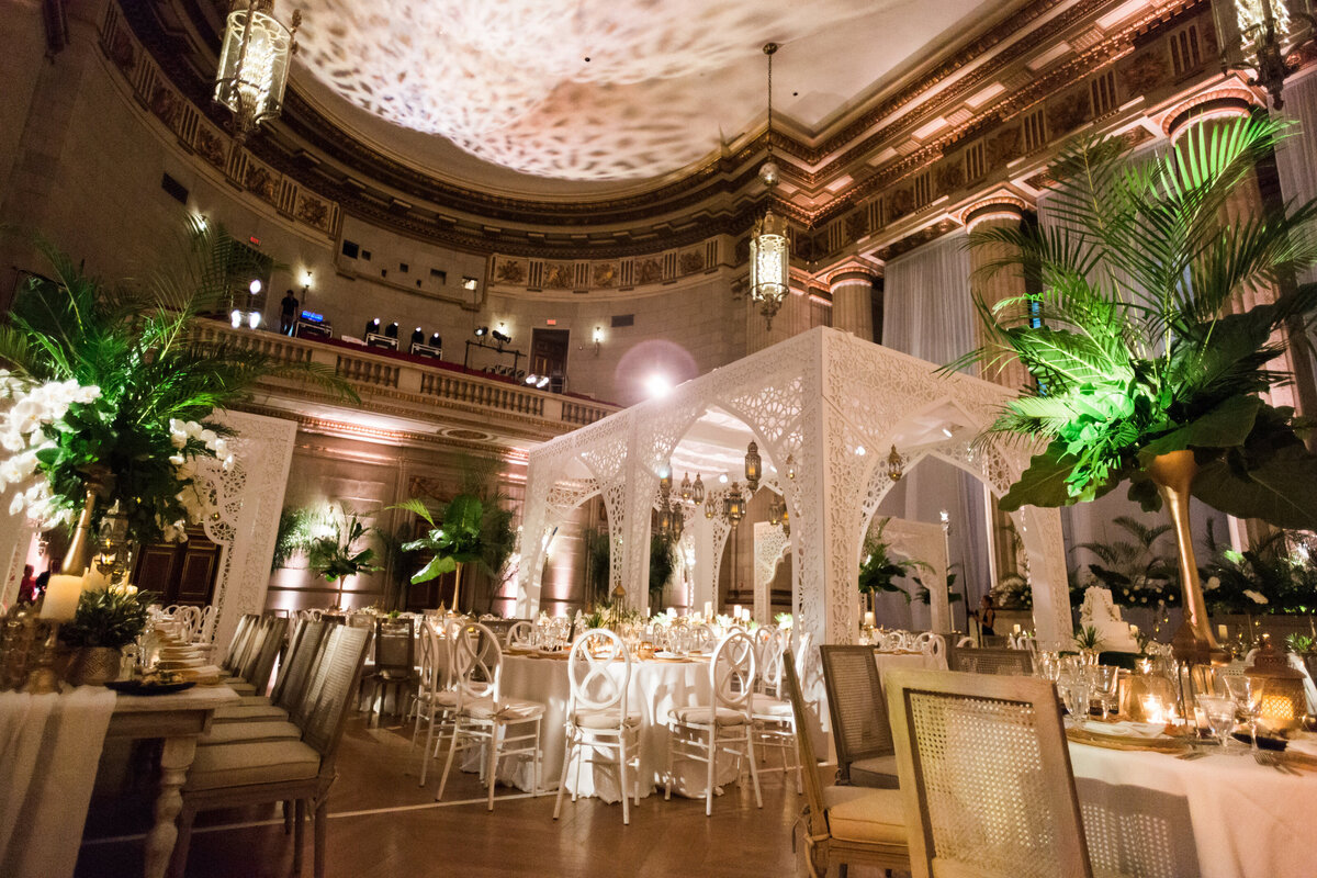 The photo capture was taken at Washington D.C., specifically at The Mellon Auditorium, showcasing one of the best luxury DC venues with European flair through the lens of photographer Sarah Bradshaw. This opulent banquet hall is ornately decorated for an event, boasting elegant white chairs and tables, grand chandeliers, lush greenery, and intricate white arches. The warm lighting enhances the sophisticated and inviting atmosphere of the venue, making it a truly extravagant setting for any special occasion.