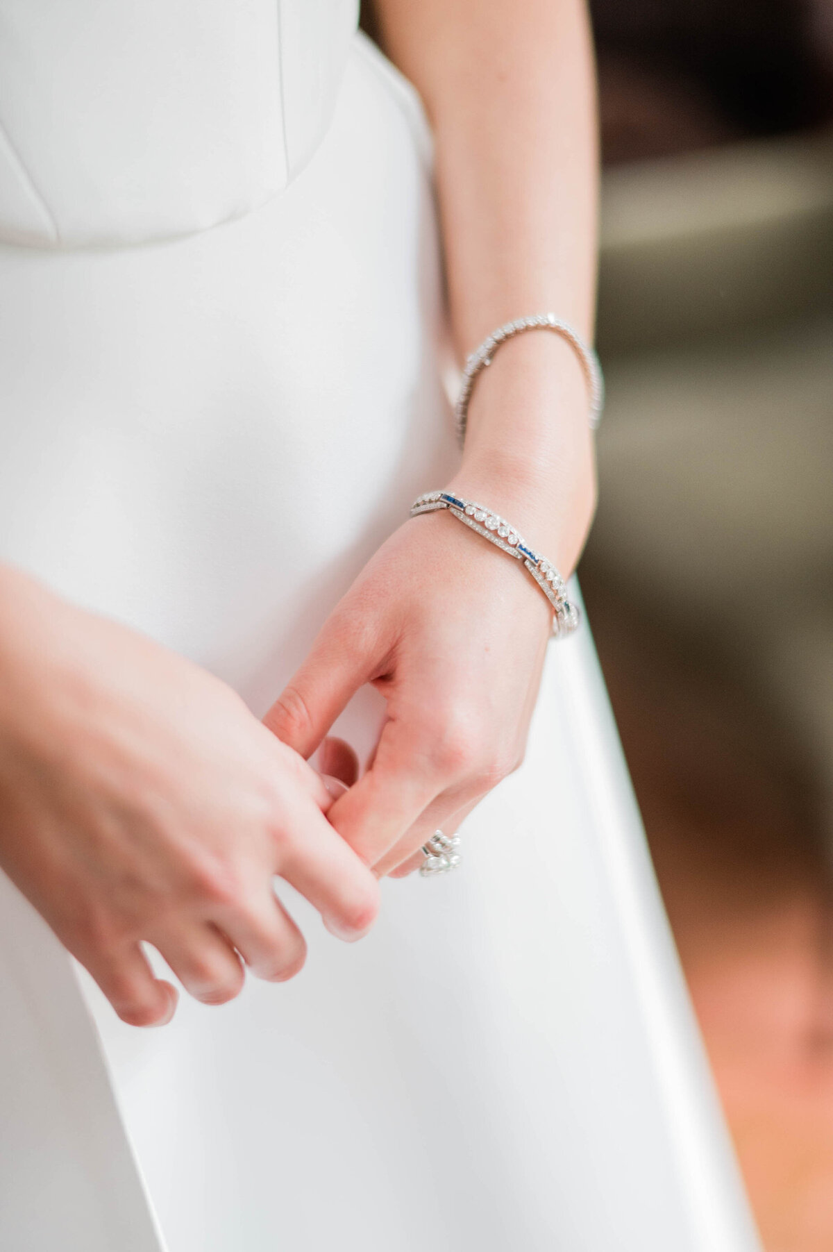 A soft-focus image of the bride’s hands, showing her delicate diamond bracelets and wedding rings against the smooth fabric of her dress.