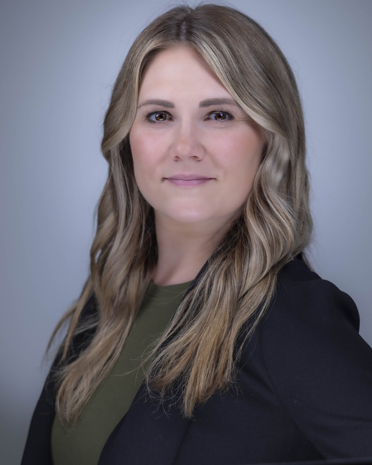 Headshot in MA of blonde woman in black blazer and green top, smiling softly against light blue backdrop by Rob Faber Photography.