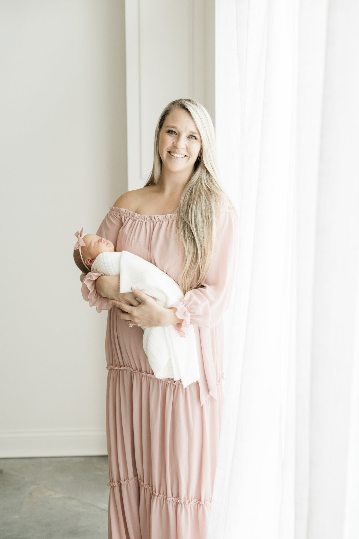Mom in soft pink tones holding her swaddled newborn in front of white curtains in a natural light studio setting.