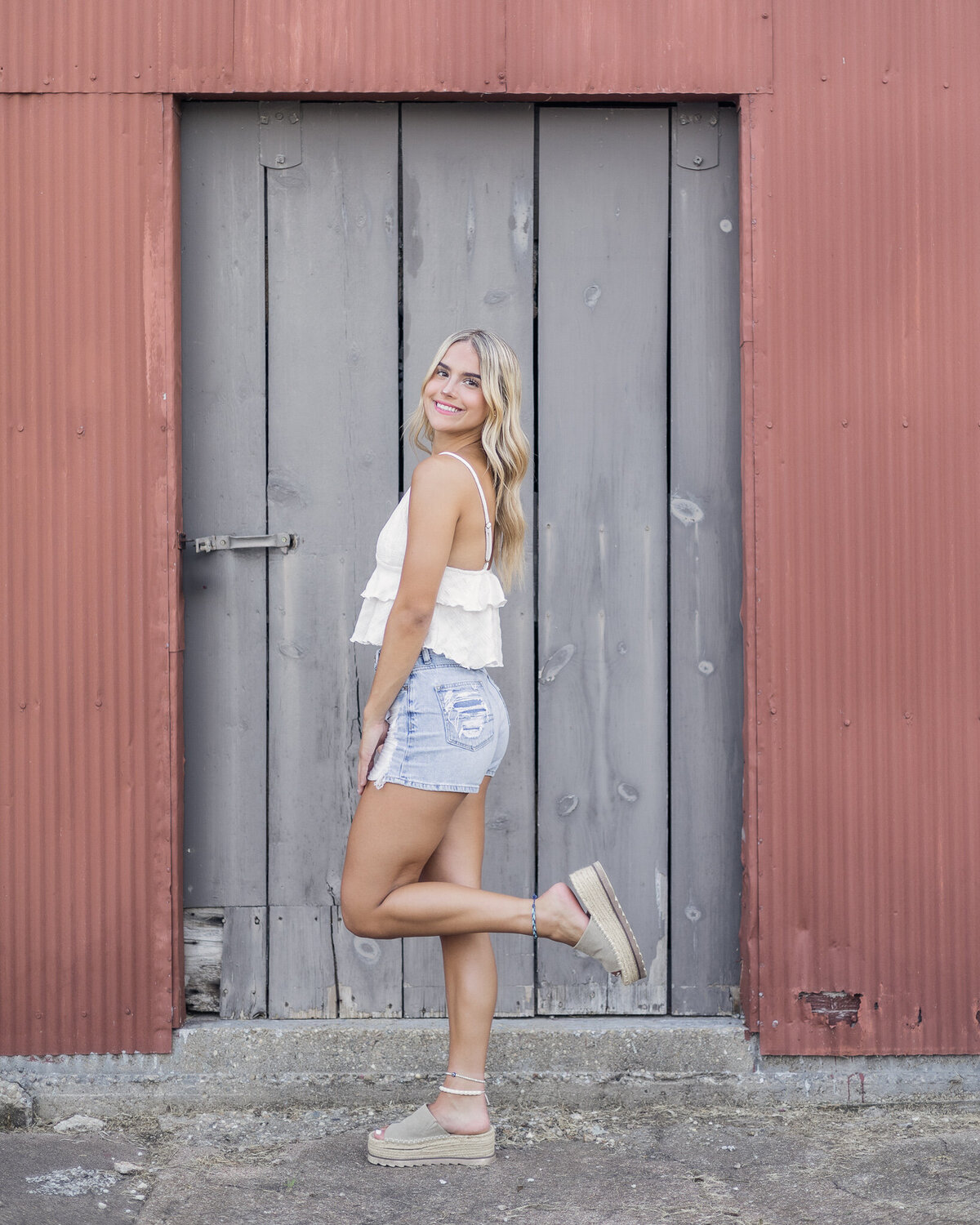 a high school female senior with long blonde hair, standing in front of a dark red building with a gray door, turned to the side with one foot up, smiling at the camera, wearing blue jeans, sandals, and white spaghetti strap shirt