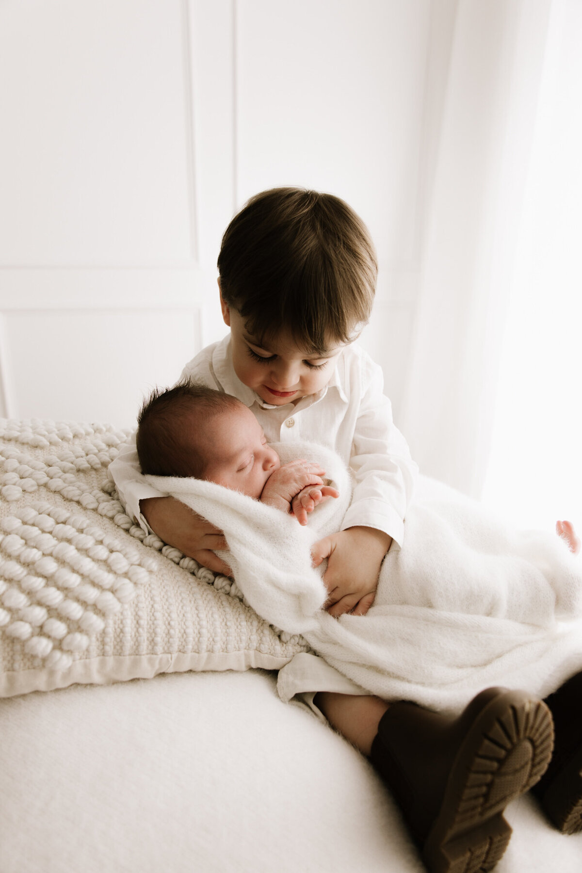 A toddler lovingly cradles their newborn sibling on a textured cream blanket, both dressed in soft neutrals, bathed in gentle diffused studio light.