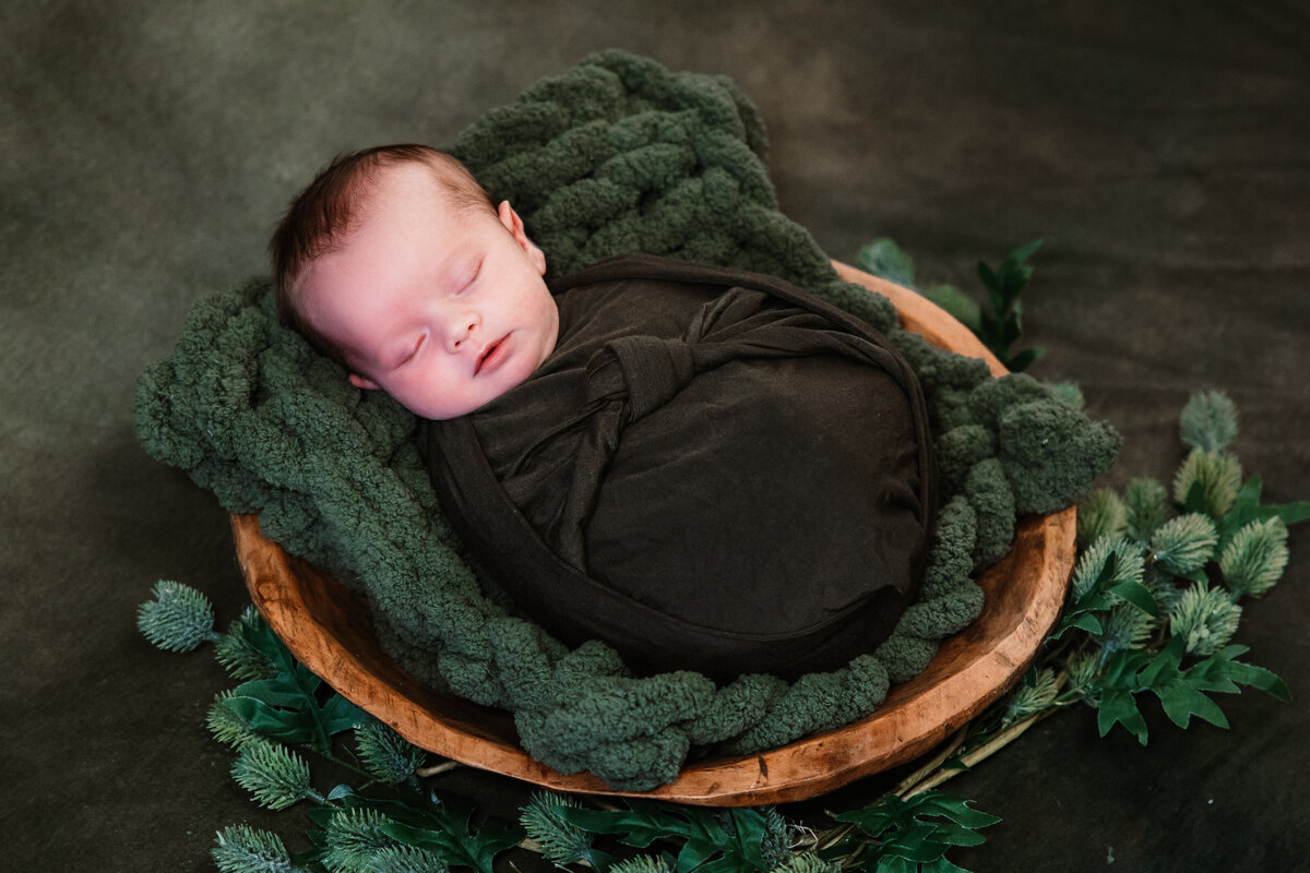 A newborn baby sleeps in a dark green swaddle positioned in a wooden bowl with greenery