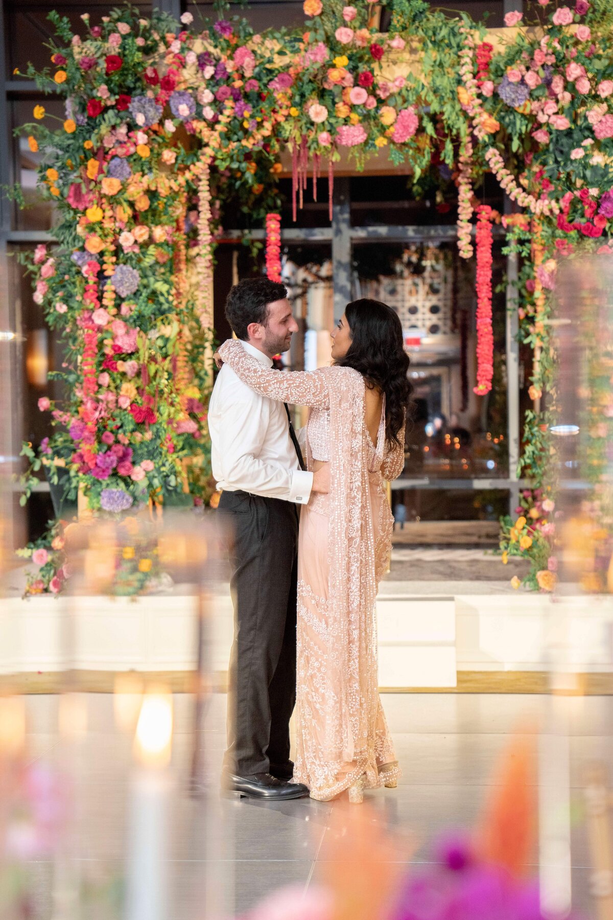 A couple dances in front of a vibrant floral arch decorated with red, pink, yellow, and purple flowers. The man wears a white shirt and black pants, while the woman wears an embellished, flowing gown. Candles are blurred in the foreground.