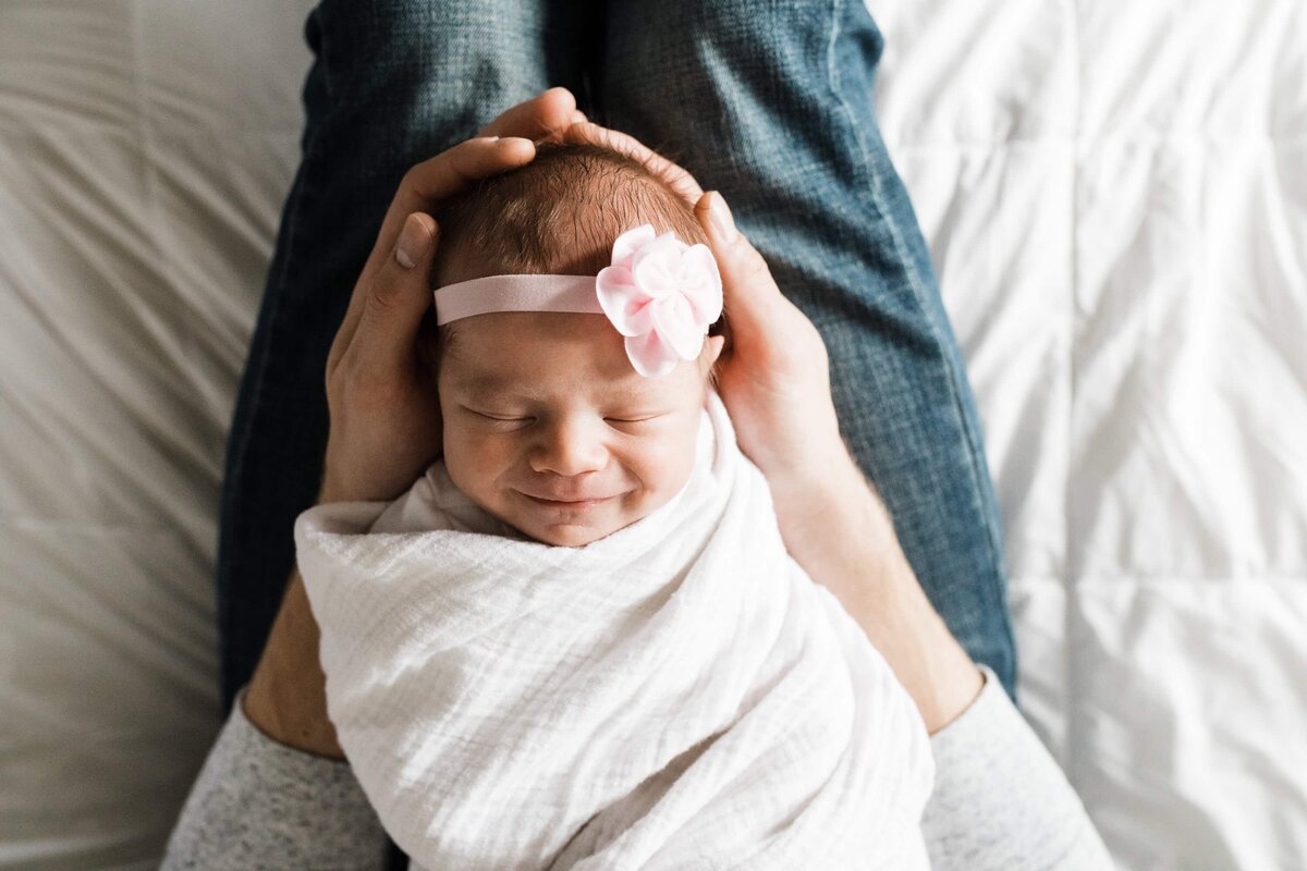 A newborn baby wrapped in a white blanket, wearing a pink headband with a flower, smiling while cradled in an adult's hands during an at-home newborn photography session.
