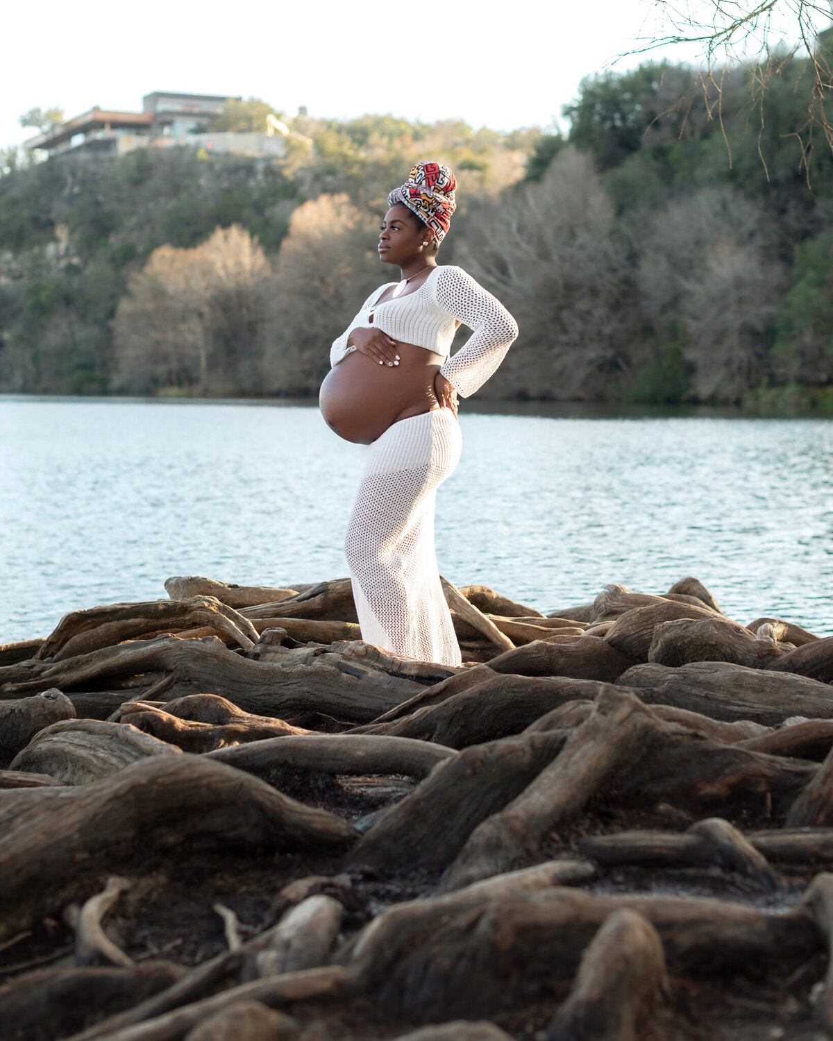 Bold maternity photo in Austin of a woman posing powerfully on twisted tree roots by the water, wearing a white crochet dress and patterned headwrap.