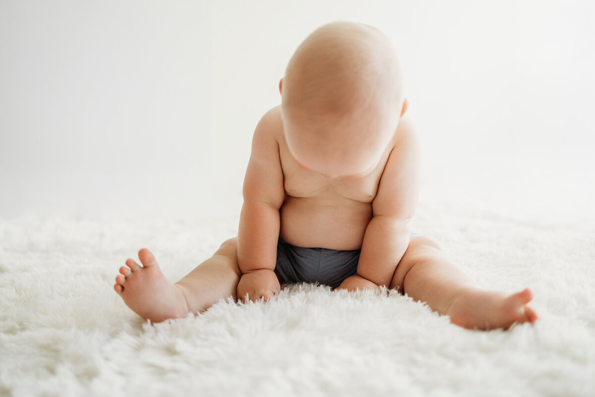 Happy six month old infant baby looking down at the fuzzy white rug while getting his photos taken for his milestone session in Denver.