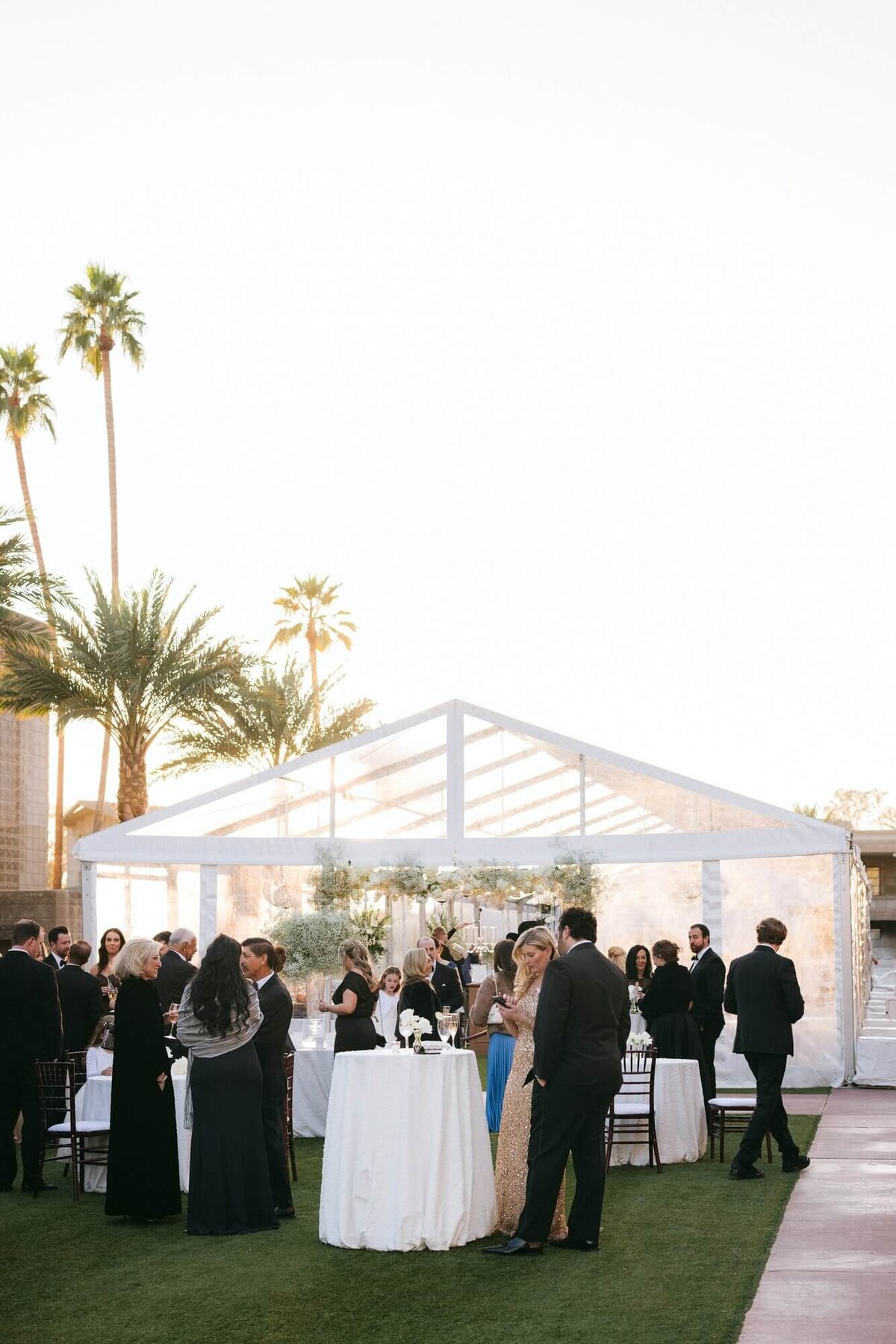Guests mingling during cocktail hour under a clear tent at the Arizona Biltmore Wedding Venue, with elegant décor and palm trees in the background.