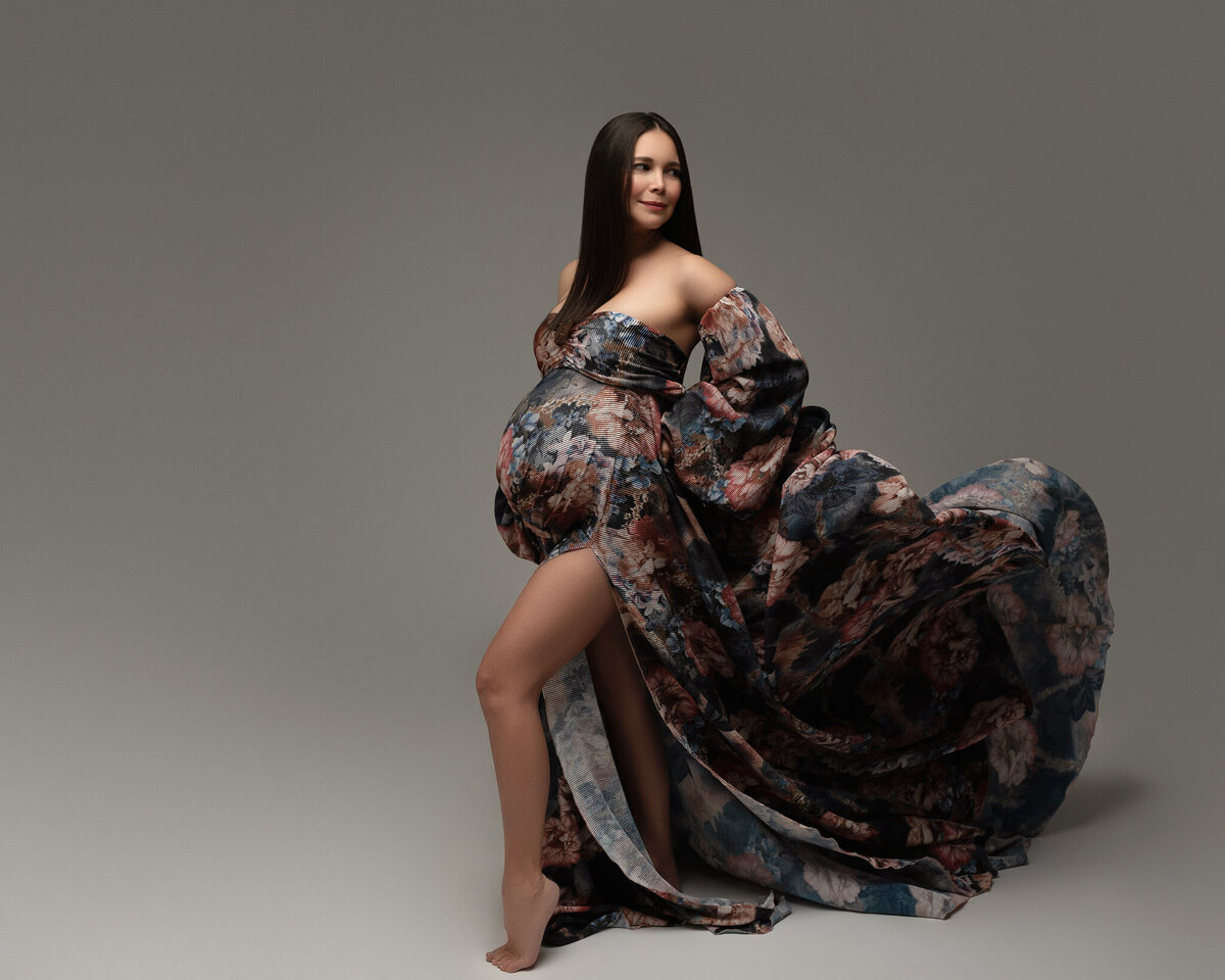 Elegant maternity portrait in a dramatic floral gown with flowing fabric, photographed in studio in Austin, Texas.