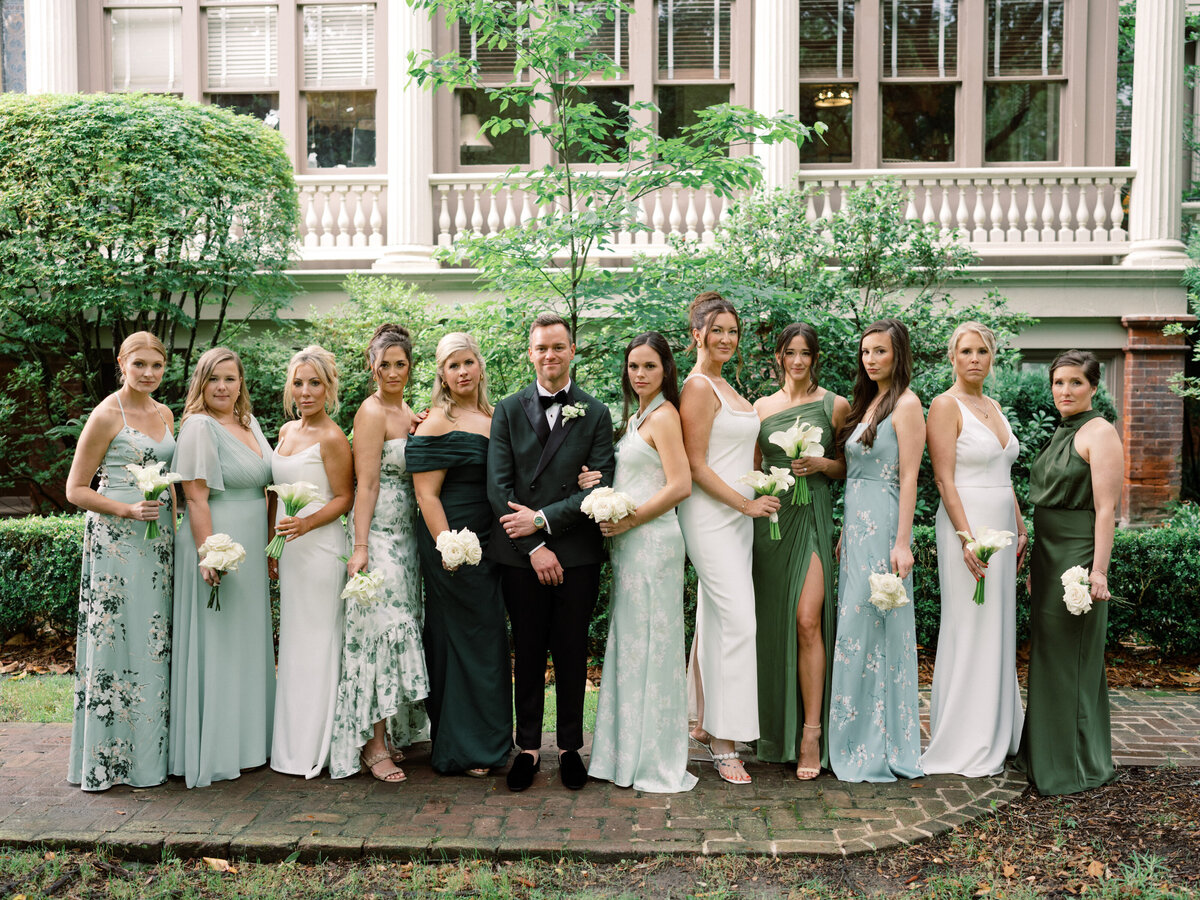 A stunning wedding party featuring the couple flanked by bridesmaids in pastel dresses engaged in a joyful celebration amidst a lush green backdrop, showcasing love and friendship.
