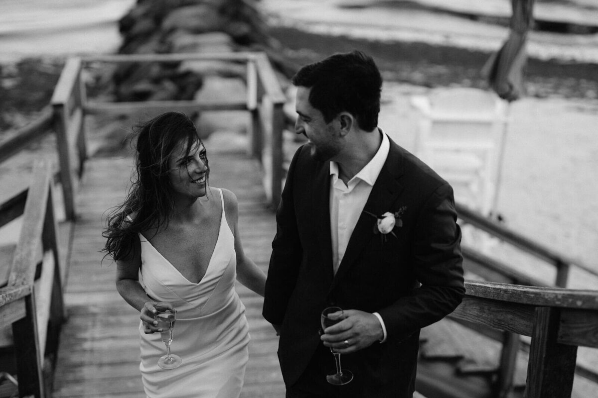 Black and white photo of the bride and the groom holding champagne glasses on a wooden platform on a seashore in Cape Cod, MA.