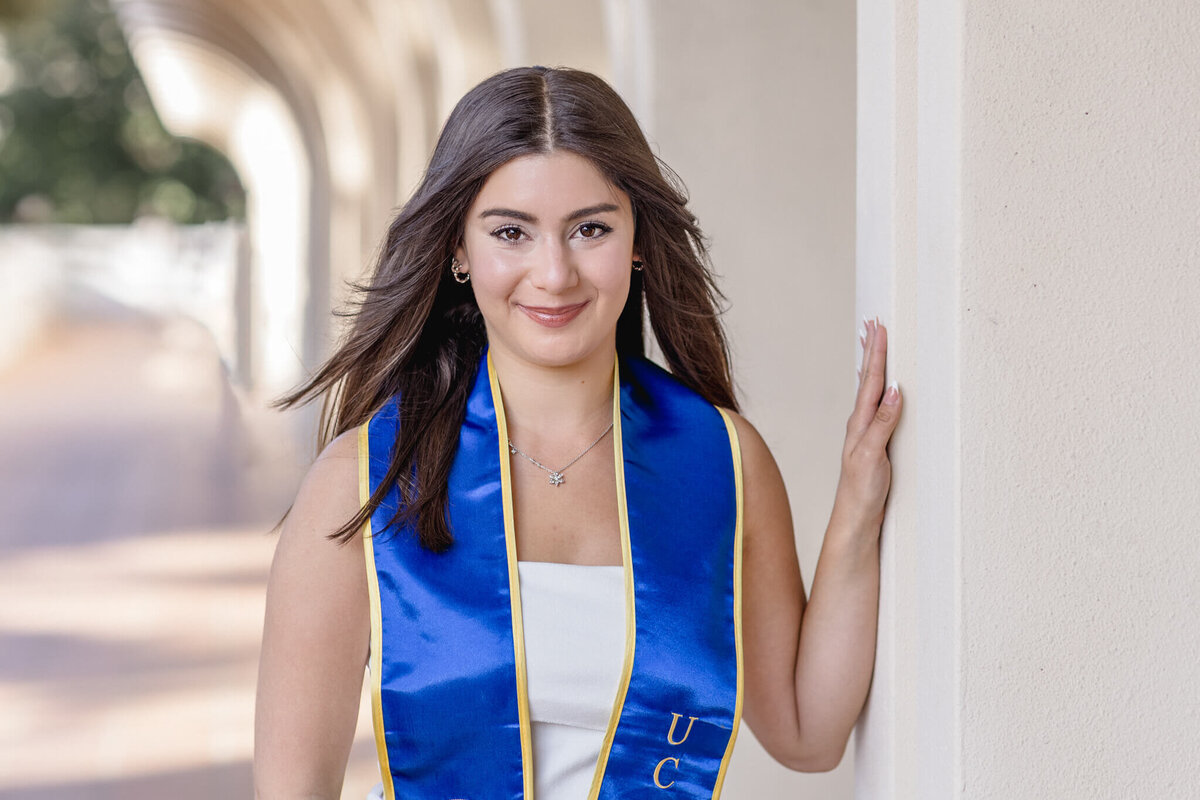 A woman with long brown hair is smiling, wearing a white dress and a blue graduation stole. She is standing outdoors, leaning against a light-colored wall with an archway in the background.