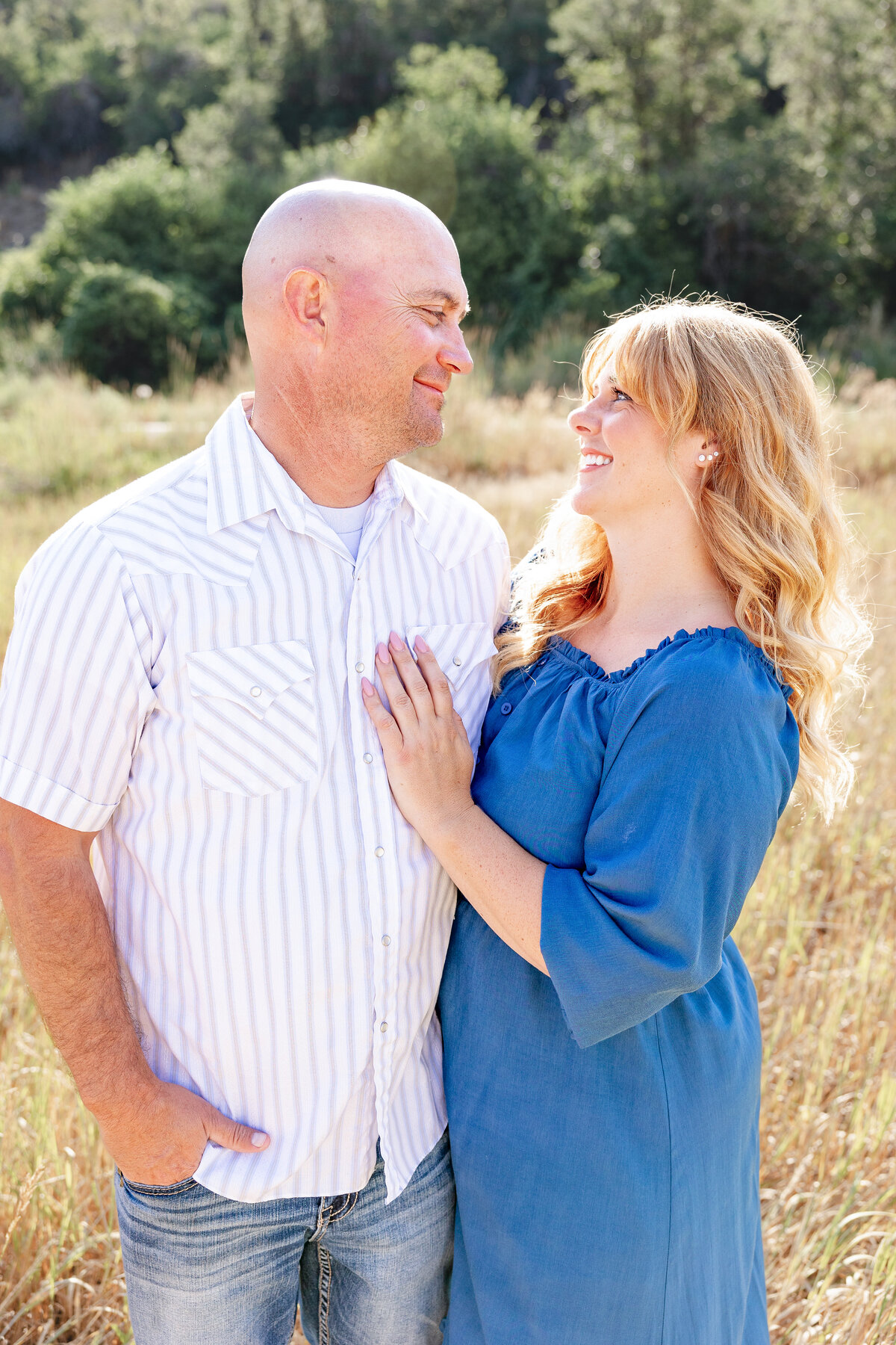 A couple gazing into each other's eyes and smiling, standing close together in a sunlit grassy field.
