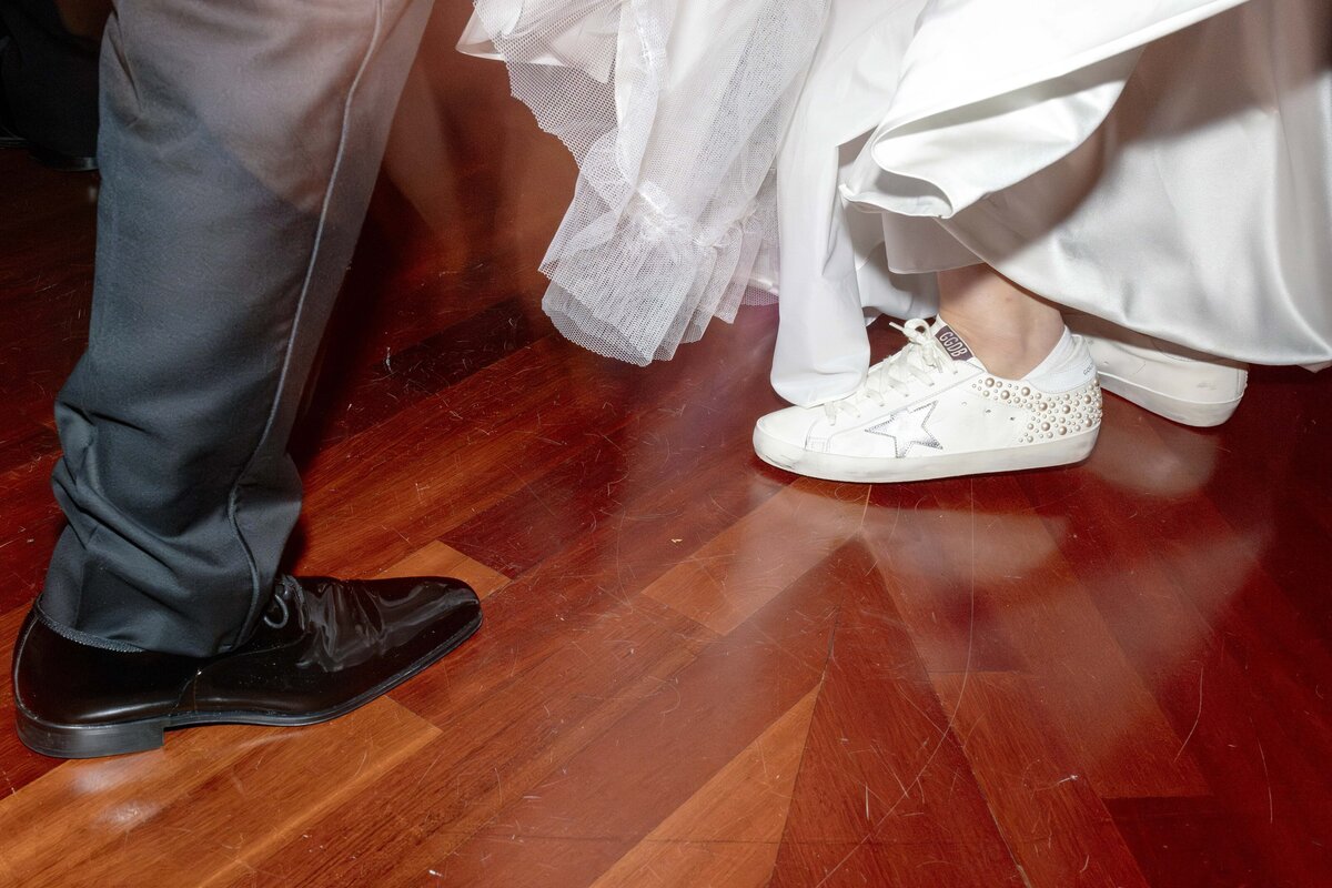 A couple dances on a wooden floor. The person on the left wears formal black shoes, while the person on the right wears white sneakers with embellishments, under a white dress.
