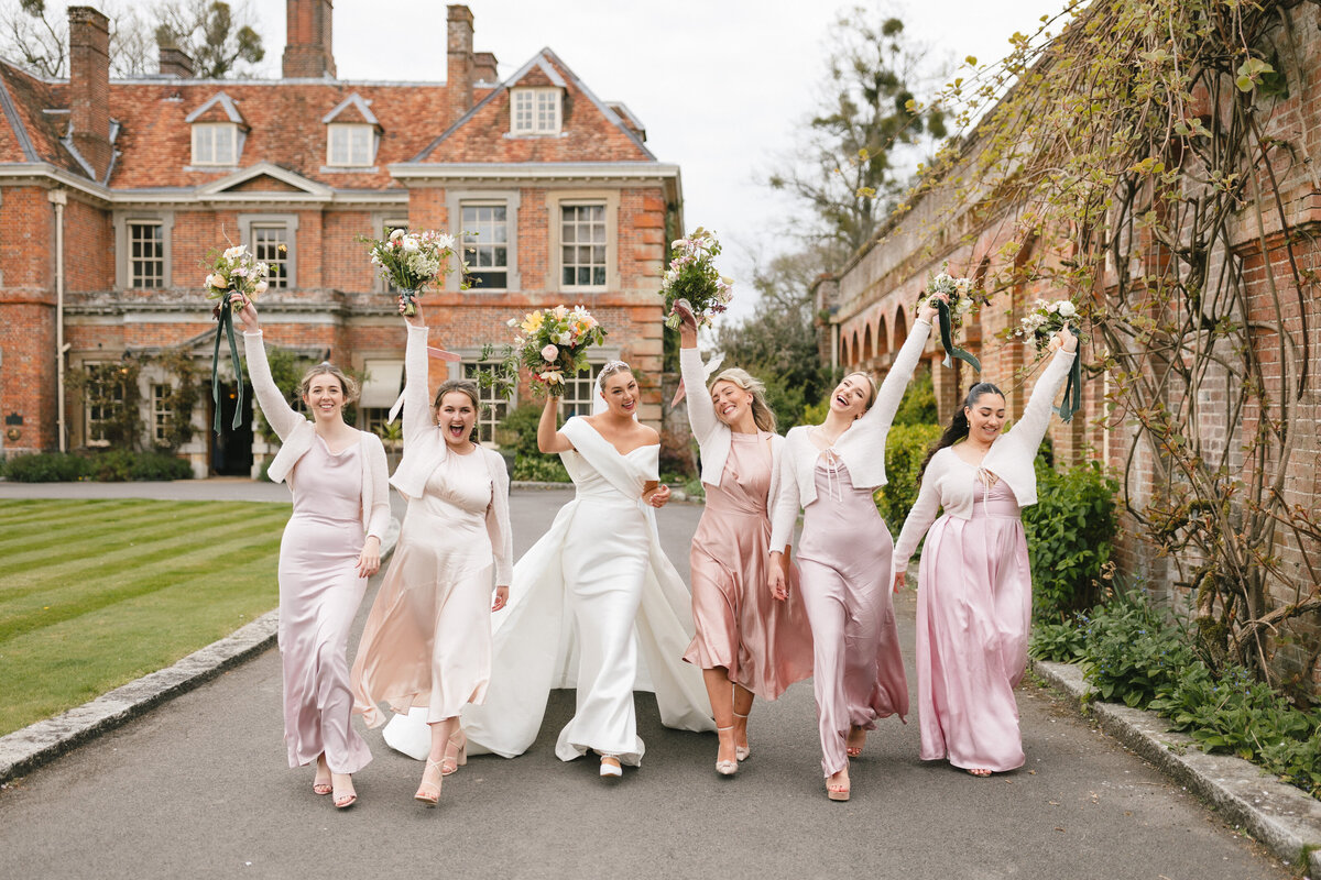 A joyful bride walks arm-in-arm with bridesmaids, all wearing pastel pink dresses, outside a large brick building. They hold bouquets aloft, celebrating in a garden setting at Lainston House