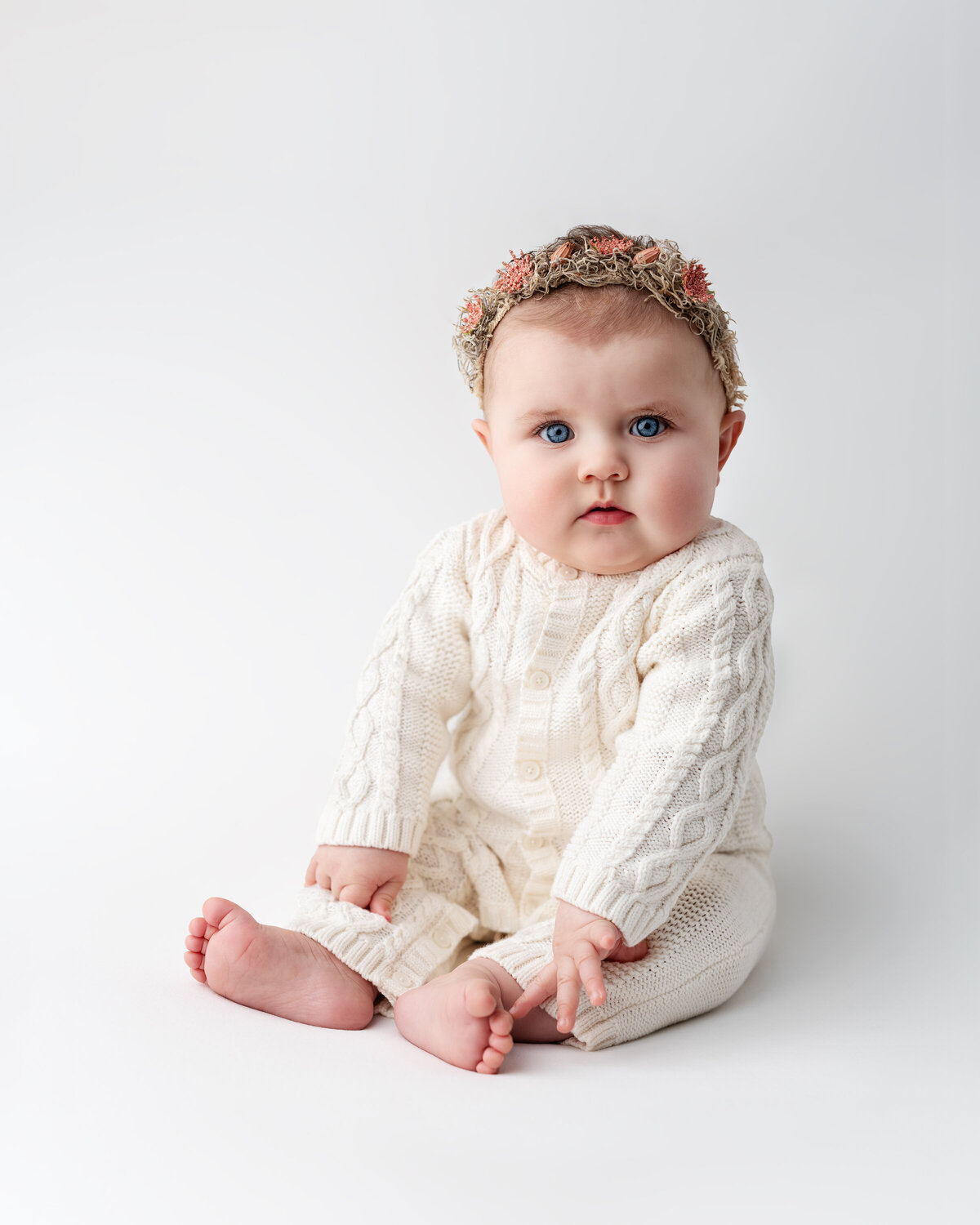 Studio milestone photo of a baby girl in a white cable-knit outfit and floral headband—timeless and softly styled