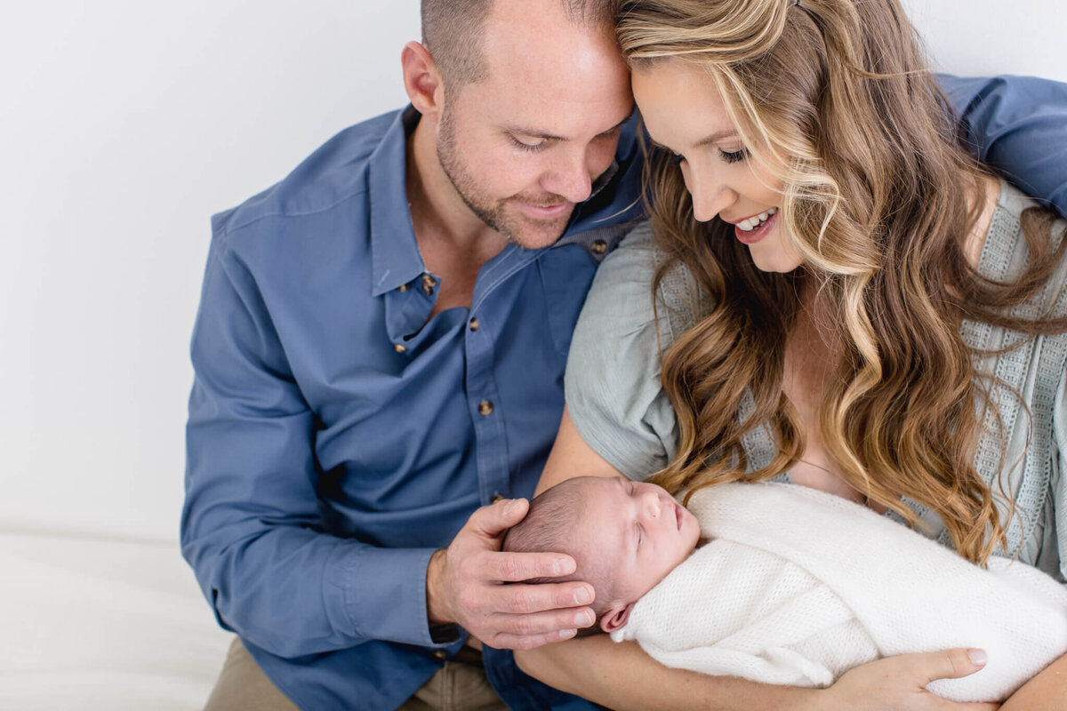 A smiling couple sits closely together, lovingly holding a newborn baby wrapped in a soft white blanket. The man is wearing a blue shirt, and the woman has long, wavy hair and is dressed in light-colored clothing.