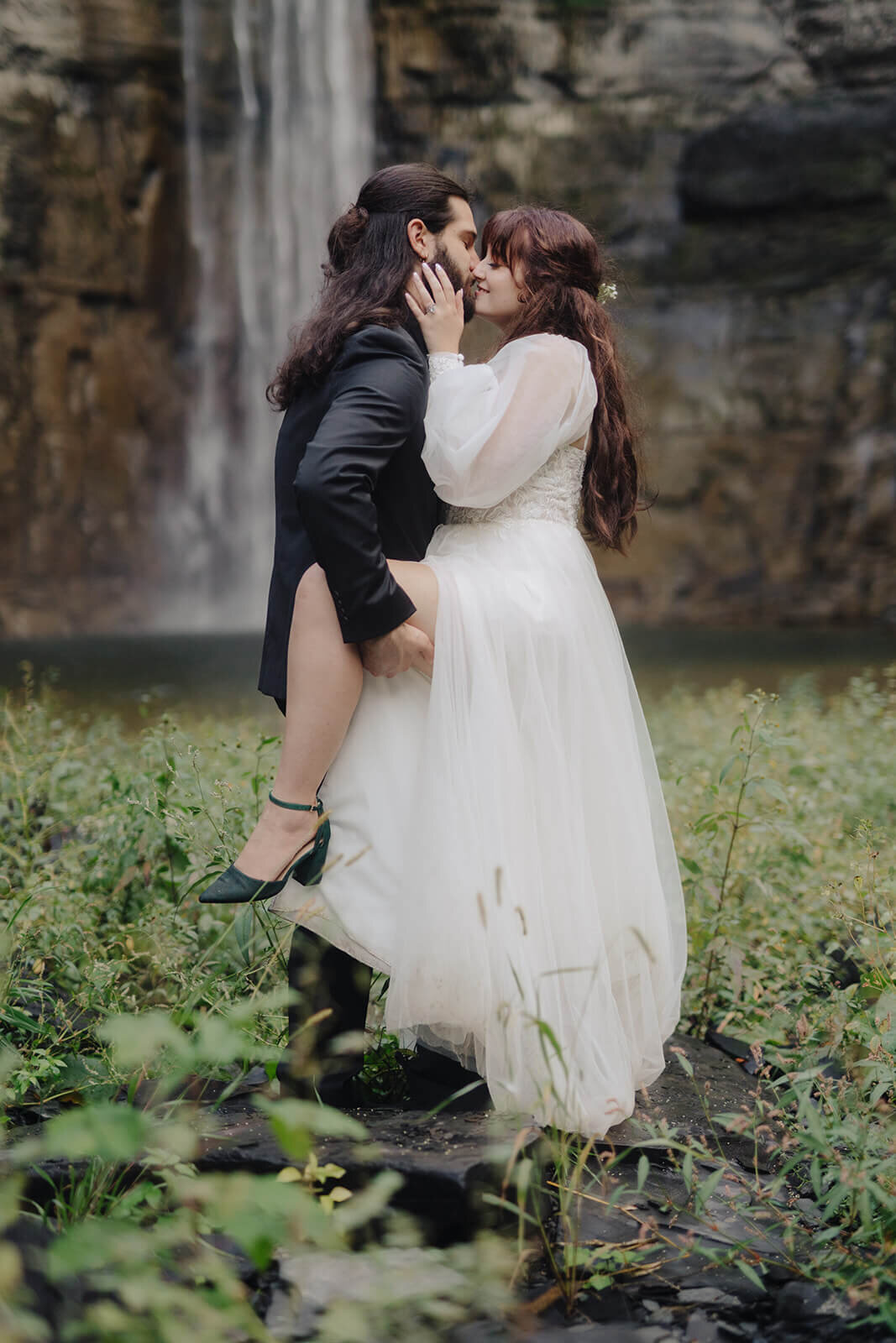 Bride and groom kissing in front of waterfall with groom lifting brides leg up