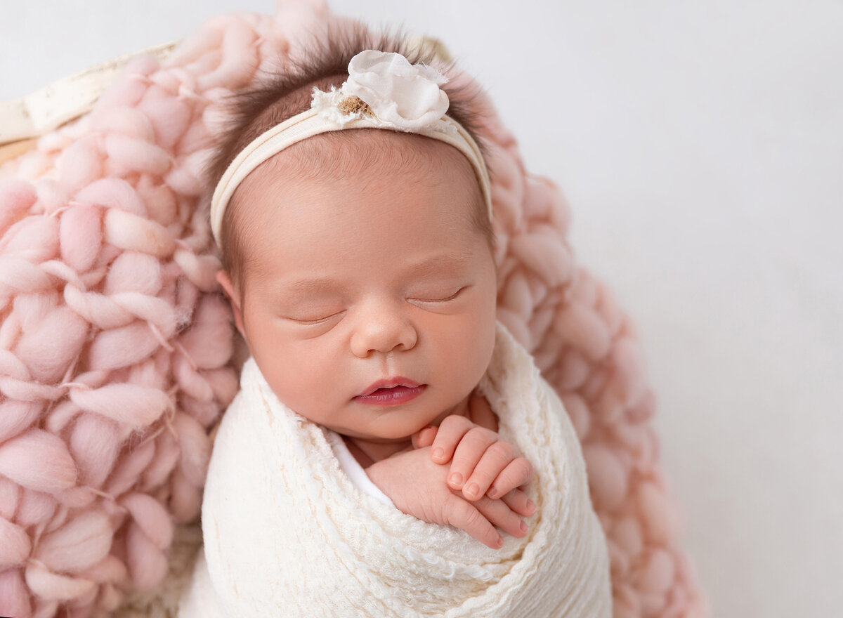 Sleeping baby girl poses for Brooklyn, NY photoshoot. Baby girl is wrapped in a cream swaddle with her hands peeking out top and resting under her chin. Baby is lying on a pink blanket.