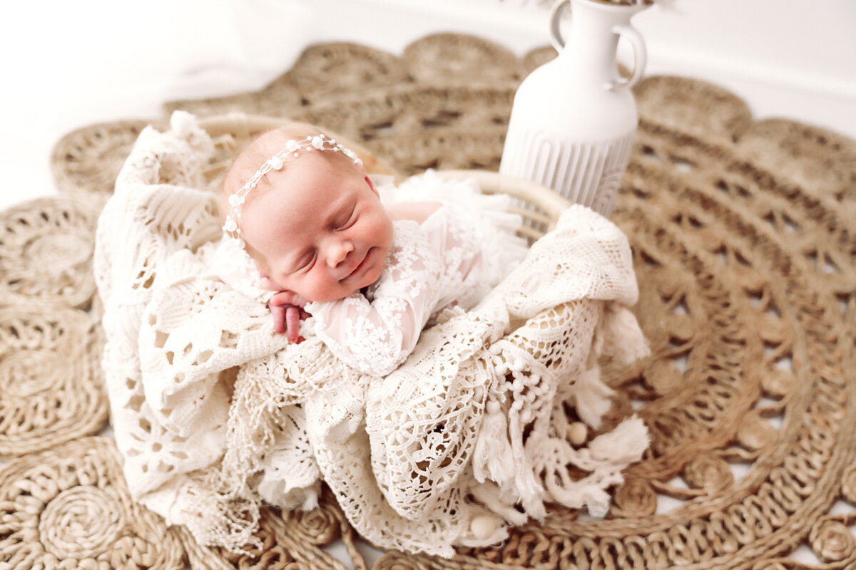 A serene newborn baby sleeps peacefully wrapped in a delicate lace blanket, adorned with a dainty headband, nestled on a woven mat, evoking a sense of warmth and innocence. Taken by Twin Cities Newborn Photographer, Fig and Olive Photography