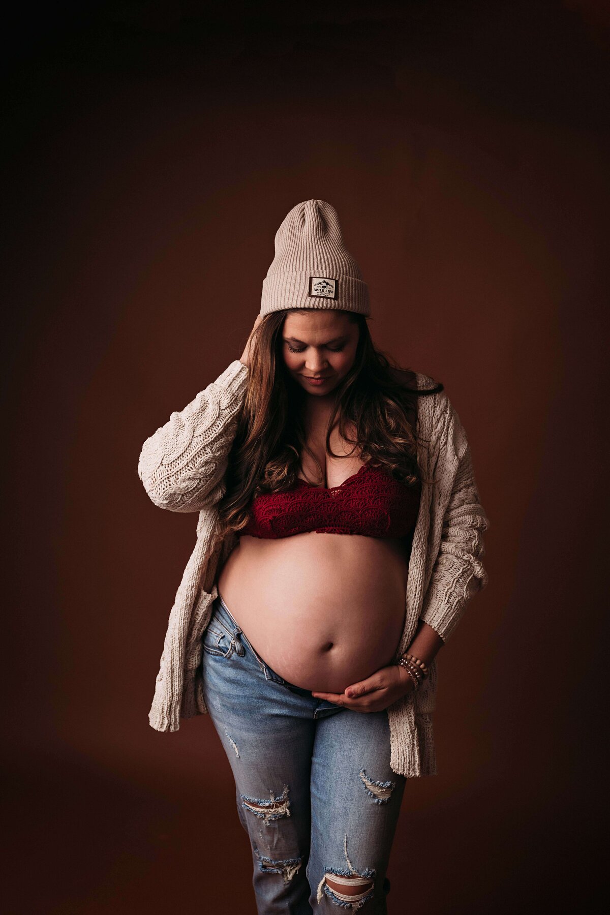 Hipster maternity portrait of a mom in blue jeans, red bra, and a beanie, posing with a cardigan draped over her shoulders. Captured in Mobile, AL