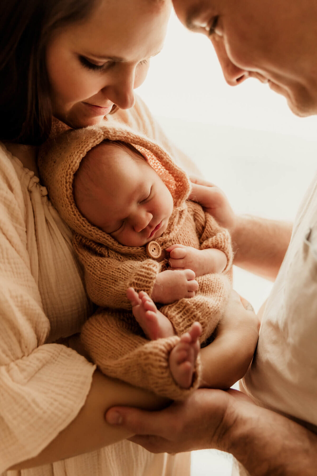 Mother and father hold their sleeping baby wearing a brown knit hooded romper.