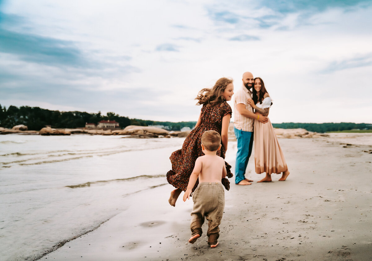 Family Photographer, mom and dad embrace as kids run on the beach