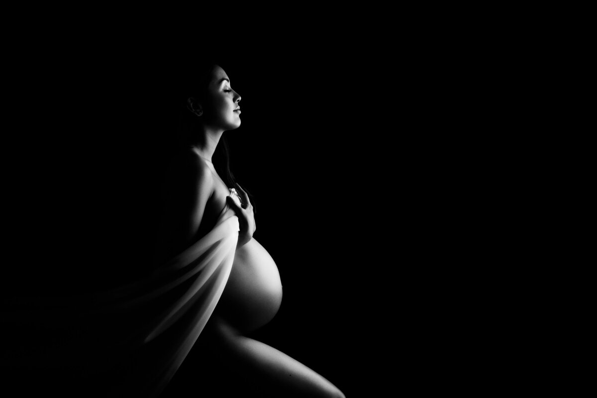 Fine art maternity portrait with a white fabric held over a woman's breasts on a black background.